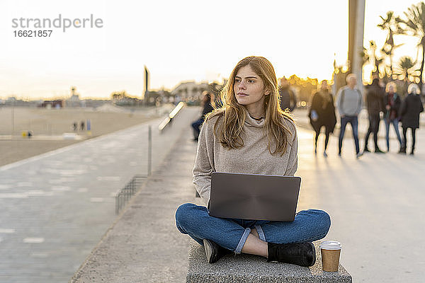 Nachdenkliche junge Frau  die im Schneidersitz mit Laptop und Einwegbecher auf einer Bank an der Promenade sitzt und wegschaut