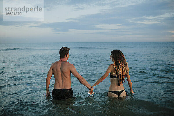 Paar in Badekleidung hält sich an der Hand  während es im Wasser am Strand steht