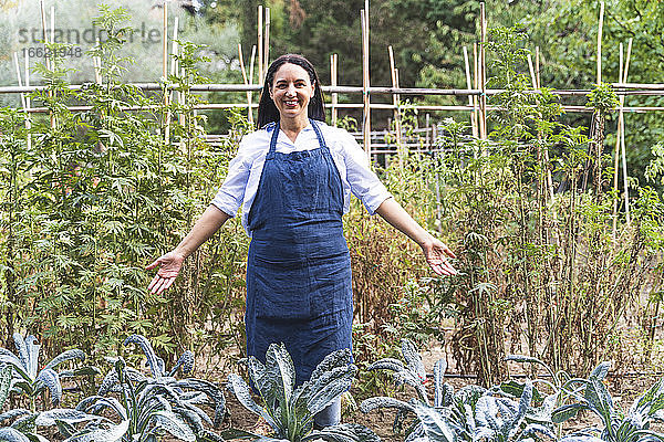 Lächelnde reife Frau mit ausgestreckten Armen  die neben frischen Bio-Kohlpflanzen im Gemüsegarten steht