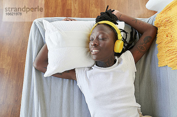 Junge Frau mit geschlossenen Augen hört Musik über Kopfhörer  während sie auf dem Sofa im Wohnzimmer liegt