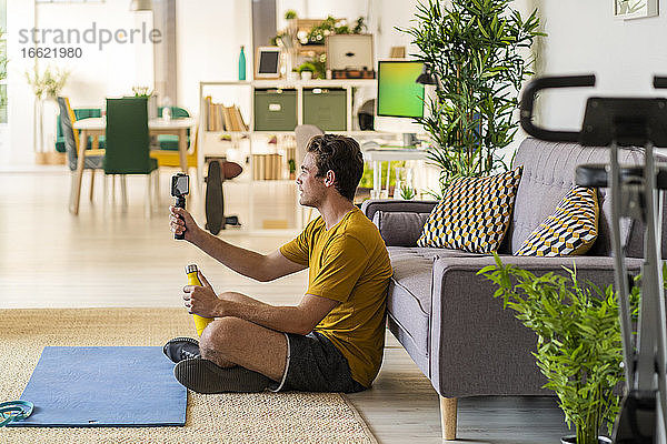 Fitnesstrainer  der ein Stativ mit Mobiltelefon hält  während er zu Hause sitzt