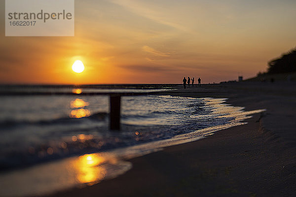 Silhouetten von vier Menschen stehen auf sandigen Küstenstrand bei stimmungsvollen Sonnenaufgang