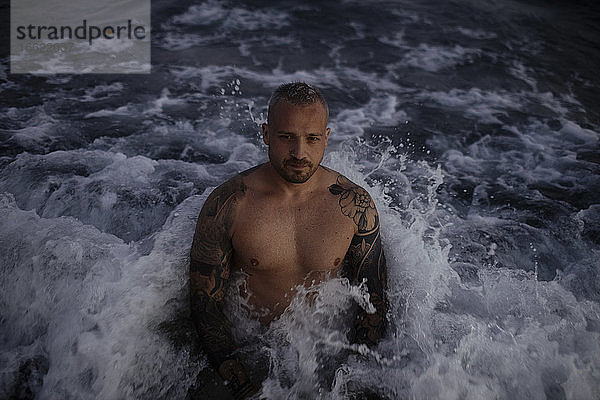 Hemdsärmeliger gutaussehender erwachsener Mann inmitten von Wellen im Meer stehend