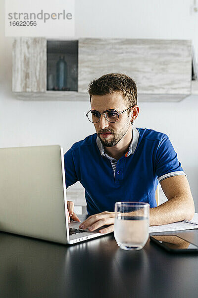 Gut aussehender junger Mann  der einen Laptop benutzt und am Tisch Hausaufgaben macht