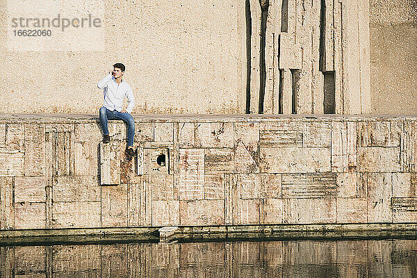 Gut aussehender junger Mann  der mit seinem Mobiltelefon spricht  während er an einem sonnigen Tag auf einer Mauer über einem Kanal sitzt