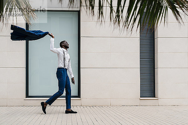 Glücklicher männlicher Unternehmer  der einen blauen Anzug hält  während er in der Stadt auf dem Bürgersteig geht