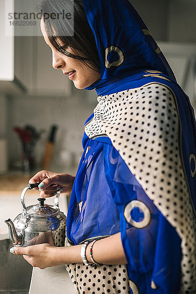 Junge Frau  die eine Teekanne hält  während sie in der Küche zu Hause steht