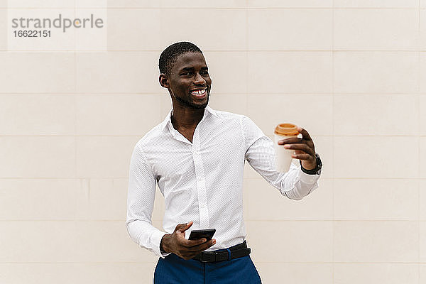 Lächelnder junger Geschäftsmann  der wegschaut  während er einen wiederverwendbaren Becher und ein Smartphone an eine Wand in der Stadt hält