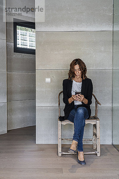 Frau  die ein Mobiltelefon benutzt  während sie auf einem Stuhl an der Wand im Büro sitzt