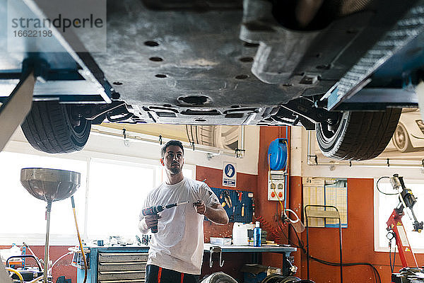 Junger Mechaniker hält Arbeitswerkzeug und betrachtet ein Auto  während er in einer Autowerkstatt steht