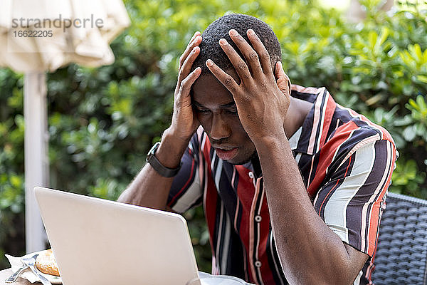 Angespannter junger Mann sitzt mit dem Kopf in den Händen und schaut auf einen Laptop in einem Café