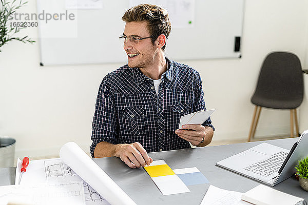 Lächelnder Mann wählt Farbmuster aus  während er am Schreibtisch im Büro sitzt