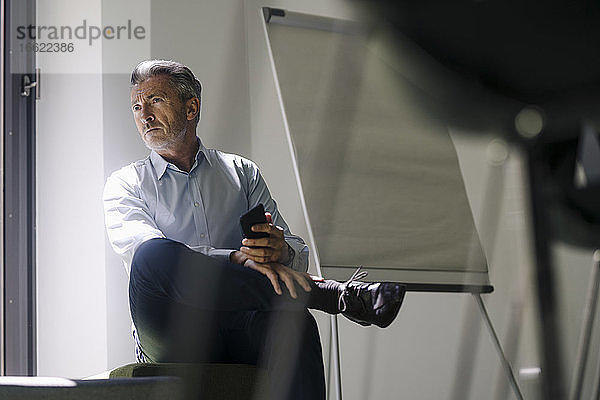 Geschäftsmann  der ein Mobiltelefon hält  während er im Büro am Fenster sitzt