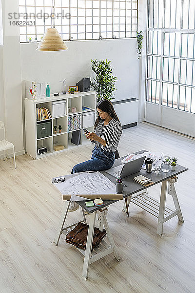 Junge Geschäftsfrau  die ein Smartphone benutzt  während sie an einem Schreibtisch mit einer Blaupause sitzt  in einem kreativen Arbeitsumfeld