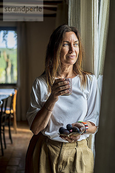 Nachdenkliche Ernährungsberaterin  die ein Getränk und Obst in der Hand hält und durch ein Fenster schaut