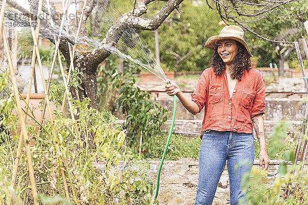 Lächelnde schöne reife Frau bewässert Pflanzen aus dem Schlauch im Garten