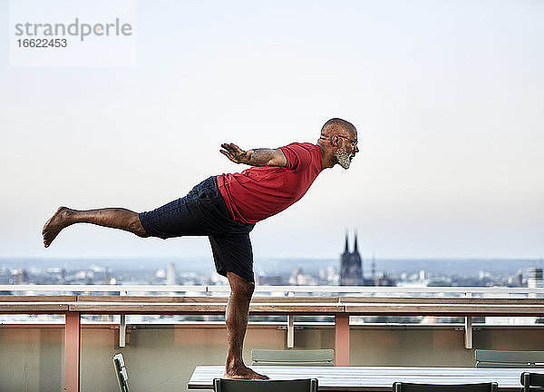 Glatzköpfiger Mann balanciert auf dem Esstisch  während er auf der Terrasse eines Gebäudes bei Sonnenuntergang Yoga übt