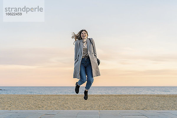 Verspielte junge Frau springt auf der Promenade am Strand gegen den Himmel bei Sonnenuntergang