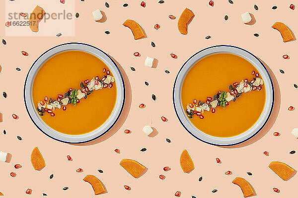 Studioaufnahme von zwei Schalen mit Kürbissuppe  umgeben von Kürbiskernen  Granatapfelkernen und Stücken von Käse und Kürbis
