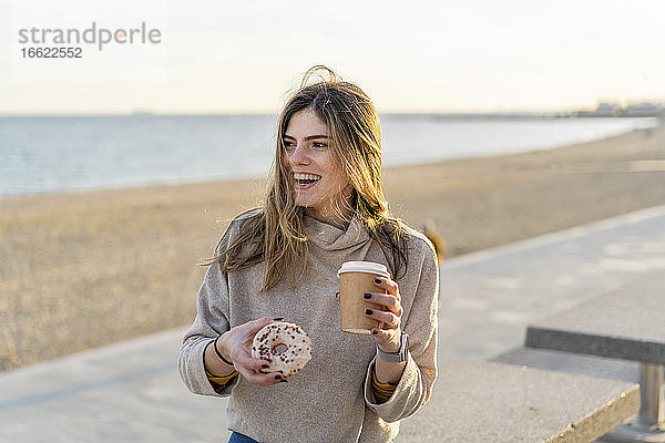 Fröhliche Frau  die einen frischen Donut mit einem Einwegbecher in der Hand hält  während sie am Strand sitzt und bei Sonnenuntergang in den Himmel schaut