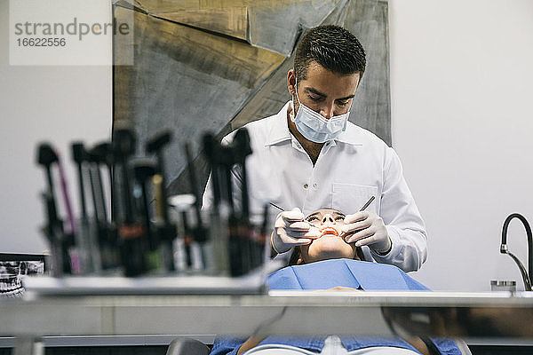 Männlicher Zahnarzt mit Maske untersucht die Zähne einer jungen Patientin während eines Besuchs in der Praxis