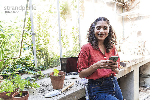Lächelnde reife Frau  die eine Textnachricht auf einem Smartphone schreibt  während sie im Gartenhaus steht