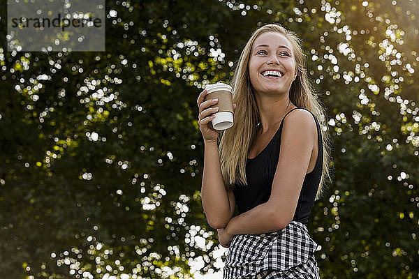 Fröhliche junge Frau hält eine Kaffeetasse in der Hand  während sie im Park vor Bäumen steht