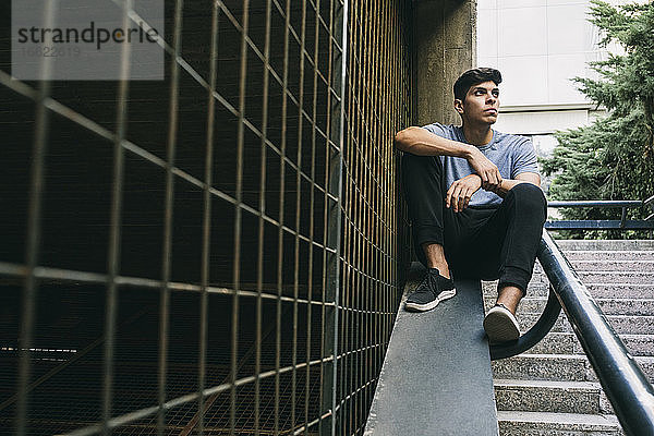 Nachdenklicher junger Mann sitzt auf einer Stützmauer am Zaun