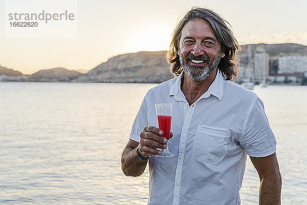 Lächelnder reifer Mann  der ein Getränk hält  während er bei Sonnenuntergang am Meer steht