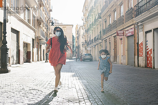 Fröhliche Mutter und Tochter mit Masken laufen auf der Straße in der Stadt