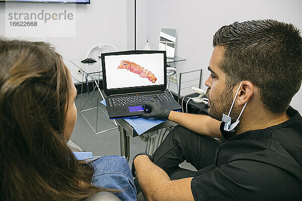 Männlicher Arzt zeigt einer Patientin ein Bild des Zahnfleisches auf einem Laptop-Bildschirm in einer Klinik