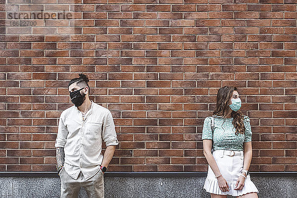 Trauriges Paar mit Gesichtsmasken  das während der Coronavirus-Krise an einer Mauer steht