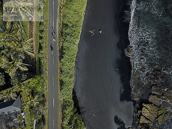 Indonesien  Bali  Luftaufnahme von Männern  die auf einer asphaltierten Straße entlang eines schwarzen Küstenstrands Skateboard fahren