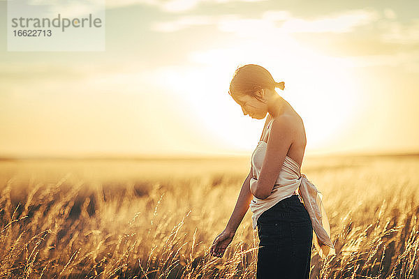 Junge Frau mit schulterfreiem Oberteil steht in einem Feld gegen den Himmel bei Sonnenuntergang
