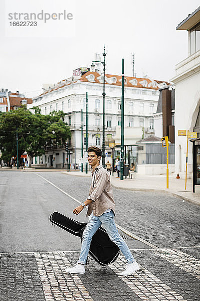 Lächelnder Mann mit Gitarre beim Überqueren der Straße in der Stadt