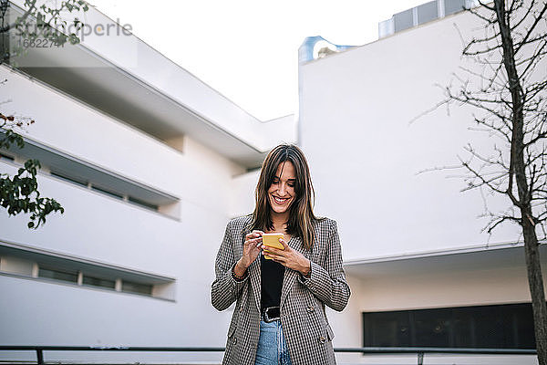 Lächelnde Frau  die eine Textnachricht auf ihrem Smartphone sendet  während sie an der Außenseite eines Gebäudes steht