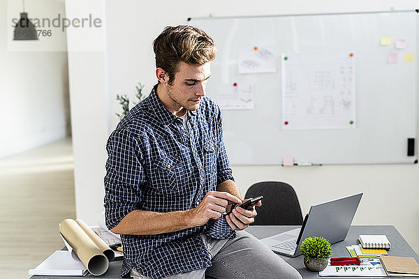 Junger Mann  der ein Mobiltelefon benutzt  während er auf dem Schreibtisch im Büro sitzt