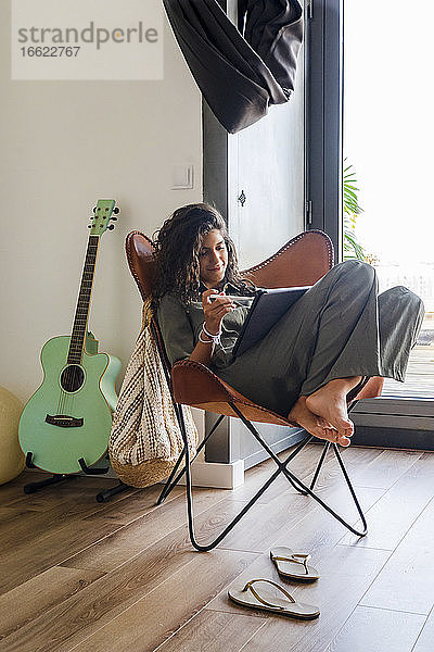 Mädchen  das ein digitales Tablet benutzt  während es zu Hause auf einem Stuhl neben einer Gitarre sitzt
