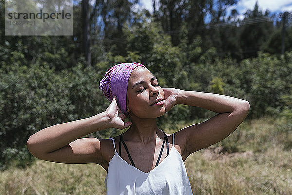 Junge Frau trägt lila Kopftuch  während sie mit geschlossenen Augen im Park an einem sonnigen Tag steht