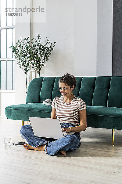 Lächelnde Geschäftsfrau  die im Schneidersitz auf dem Boden sitzt und einen Laptop auf dem Sofa im Büro benutzt