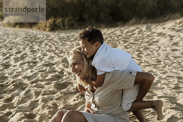 Enkel  der seine Großmutter von hinten umarmt  während er im Sand am Strand sitzt