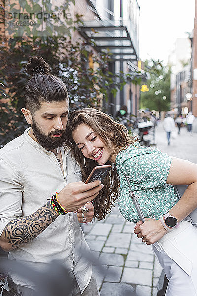 Hipster-Mann zeigt seiner Freundin sein Smartphone  während er auf dem Gehweg steht