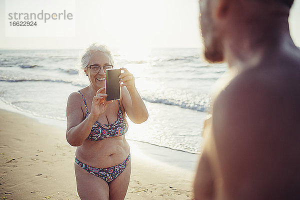 Ältere Frau im Bikini fotografiert einen Mann am Strand