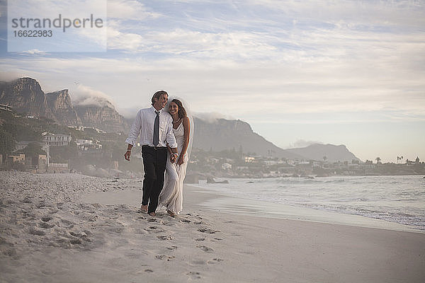 Glückliches frisch verheiratetes Paar spaziert am Strand gegen den Himmel bei Sonnenuntergang