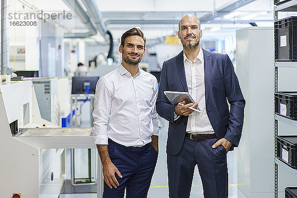 Selbstbewusster reifer Geschäftsmann  der ein digitales Tablet hält  während er neben einem jungen männlichen Ingenieur in einer Fabrik steht