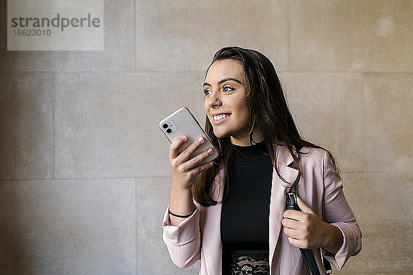 Lächelnde junge Geschäftsfrau  die mit ihrem Smartphone an der Wand eines Cafés spricht