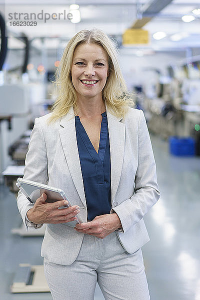 Lächelnde reife blonde Geschäftsfrau  die ein digitales Tablet hält  während sie vor einer beleuchteten Industrie steht