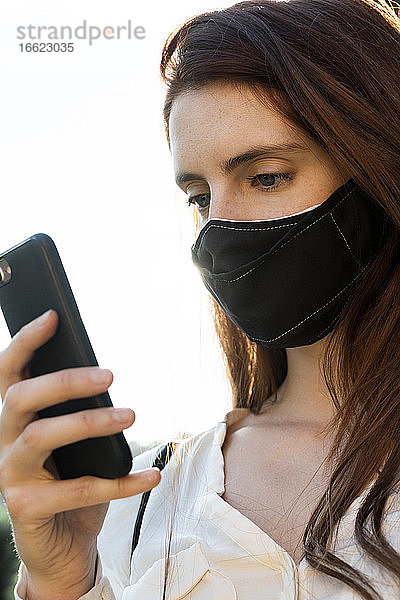 Junge Frau  die eine Textnachricht auf ihrem Smartphone schreibt und eine Gesichtsmaske trägt