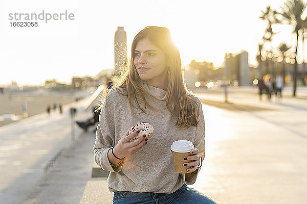 Nachdenkliche junge Frau  die einen frischen Donut mit einem Einwegbecher in der Hand hält  während sie an der Promenade sitzt und bei Sonnenuntergang wegschaut
