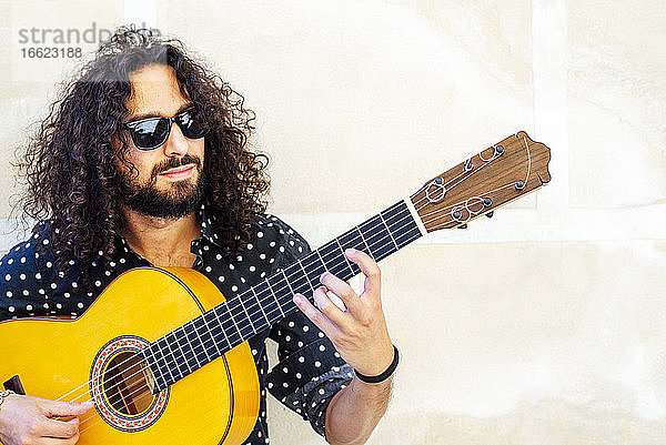 Mann mit Sonnenbrille spielt Gitarre und steht an der Wand